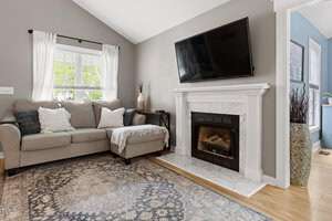 Cozy living room with a gray sofa, patterned rug, and pillows. A TV is mounted above a white fireplace, and a window with white curtains lets in natural light. A small table with a vase is beside the sofa. Walls are painted in neutral tones.