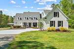 A large white two-story house on 3195 Range Road boasts a gray roof, multiple gables, and a covered front porch. The gravel driveway gracefully leads to a landscaped yard with shrubs and trees in the background. It's a bright, sunny day with just a few clouds adorning the sky.