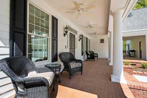 A spacious front porch with white siding and wooden flooring. It features black wicker chairs with cushions, ceiling fans, and decorative columns. The area is well-lit with outdoor wall lanterns and bordered by green foliage.