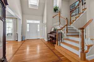 A spacious, well-lit foyer with wooden floors, white walls, and a grand staircase. A piano is positioned near the staircase, with a plant on top. Large windows and a decorative glass door let in natural light.