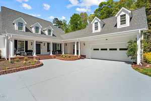 A spacious white house features a sloped roof with dormer windows, a welcoming front porch, and a two-car garage. The driveway is expansive, and there are well-maintained shrubs and trees surrounding the property.