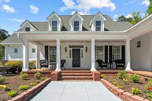 A white two-story house with a gray roof, featuring a covered front porch with brick steps. The porch has chairs and decorative columns. A landscaped garden with small plants and a decorative urn is in the foreground under a partly cloudy sky.