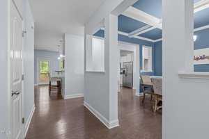A modern home interior with light blue walls and white trim. The hallway leads to a dining area and kitchen with stainless steel appliances. Wooden flooring is throughout, and a door opens to a deck outside. Hanging lights illuminate the spaces.