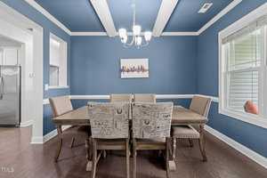 A dining room with a wooden table and six upholstered chairs, two of which have patterned fabric. The walls and ceiling are blue, with a chandelier hanging above. A small cityscape painting is on one wall, and a window is on the right.