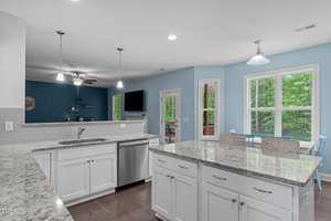 Modern kitchen with white cabinets, granite countertops, and stainless steel appliances. An island with seating faces a cozy living area with a blue accent wall and wall-mounted TV. Large windows and glass doors offer views of lush greenery outside.