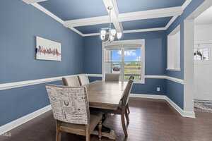 Dining room with blue walls and white trim, featuring a wooden table and chairs. One chair has printed upholstery. A chandelier hangs from a coffered ceiling, and there's a large window with a view of greenery. A cityscape painting adorns the wall.