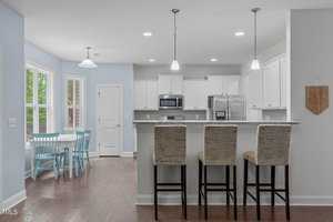 Bright, modern kitchen with white cabinets and stainless steel appliances. A marble island with three upholstered bar stools faces the kitchen. A dining area with a round table and light blue chairs is near a window, illuminating the space.