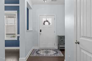 A modern, light-filled foyer with pale blue walls, a white front door with a window, and dark hardwood floors. A patterned rug lies on the floor, and a small cabinet sits near the door. Natural light illuminates the space.