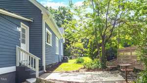 Side view of a blue house with a white trim, featuring a small patio area with two wicker chairs. The area is surrounded by greenery, with tall trees and a wooden fence providing privacy and shade. The sky is clear and sunny.