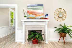 A decorated white fireplace with a colorful abstract painting above it. A potted orchid and blue vase sit on the mantel. Ferns in wicker baskets are on the hearth. A round mirror with a geometric frame hangs on the wall, reflecting natural light.
