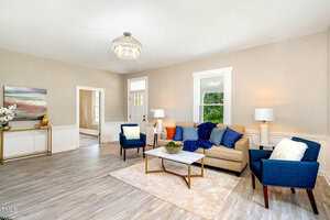 A modern living room with a beige sofa, blue armchairs, and a white marble coffee table on a light rug. There's a large window, a door, and a chandelier above. The walls are light gray, and there's a sideboard with a colorful painting.