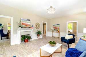A bright living room with light wood flooring, featuring a white fireplace, abstract art, and a chandelier. There's a blue armchair, a coffee table with decor, and potted plants. A glimpse of the dining area is seen through an open doorway.