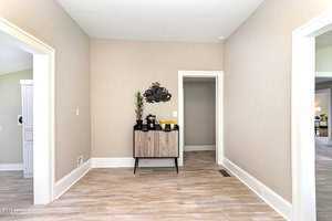 A spacious room with light wood flooring and beige walls. A small cabinet with a decorative sign and potted plant is against one wall. Doors lead to other rooms, and natural light brightens the space.