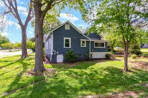 A blue house with white trim set amidst green grass and trees. The sky is bright blue with scattered clouds, and a sidewalk runs beside the lawn.