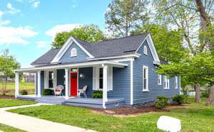 A charming blue cottage with white trim on 125 Belle Street, featuring a red front door and a covered porch with chairs. Surrounded by Oxford's lush green grass, trees, and a clear sky. A welcoming sidewalk leads to the entrance.