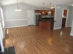 An open-concept kitchen and dining area with wood laminate flooring. The kitchen features wooden cabinets, stainless steel appliances, and a curved counter. A hanging light fixture illuminates the dining space, and several windows offer natural light.