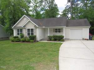 Located at 1200 E Middleton Drive, this charming single-story beige house in Creedmoor features a gabled roof, white trim, and a cozy front porch. It boasts a one-car garage, neatly trimmed bushes, and a grassy lawn with a paved driveway framed by trees.