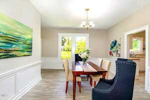 A dining room with a wooden table and beige upholstered chairs. A blue high-back chair is at the table's end. Large windows with a view of greenery and two vibrant paintings adorn the walls. A chandelier hangs above the table.