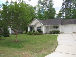 A small, single-story house with a gray roof and white siding. It has a one-car garage and a concrete driveway. There's a well-maintained lawn with shrubs and a tree in the front yard, surrounded by tall trees in the background.