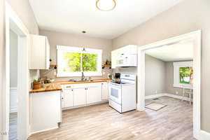 A bright kitchen with white cabinets, a wooden countertop, and a white stove. A window above the sink lets in natural light. The room has light wood flooring and opens into a sunny adjacent room with more windows.