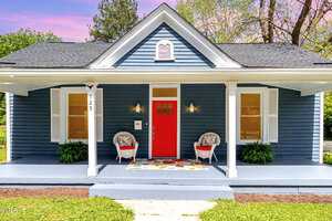 A charming blue house with white trim and a vibrant red front door. Two white chairs with colorful cushions sit on the porch, alongside a colorful doormat. The yard is landscaped with green plants, and the sky is pink and blue.
