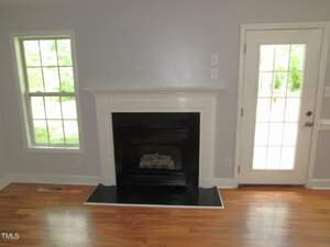 A room with light gray walls and wooden flooring features a black and white fireplace centered between two large windows. To the right, there is a glass door leading outside, allowing natural light into the space.