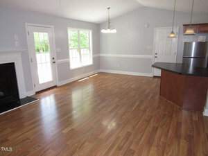 Spacious kitchen and living area with light wood flooring, light gray walls, and a high ceiling. Features a fireplace on the left, hanging lights, and large windows allowing natural light. Includes a stainless steel fridge and a door leading outside.