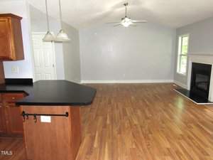 An empty living room with wood flooring, a ceiling fan, and pendant lights over a kitchen counter. Light gray walls and a large window brighten the space. A fireplace is positioned on the right side of the room.