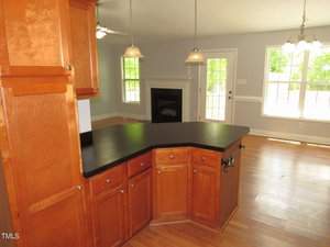 A kitchen with wooden cabinets and a black countertop, featuring an island. In the background, there's a living area with a fireplace, hardwood floors, and a ceiling fan. Large windows and glass doors allow abundant natural light.