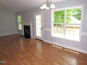 A bright, empty living room with light wood flooring. There is a fireplace, a chandelier, large windows, and a glass door leading to a deck outside. Walls are painted light gray with white trim. The room is filled with natural light.