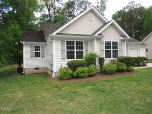 A small, single-story white house with a gabled roof and several windows. It is surrounded by a green lawn and has a few bushes planted in front. Tall trees are visible in the background.