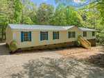 A beige mobile home with blue shutters and a metal roof sits peacefully at 3196 Loblolly Lane, Wake Forest, surrounded by lush green trees. A gravel driveway leads to the front steps, where a small path outlined with stones enhances the serene, wooded setting.