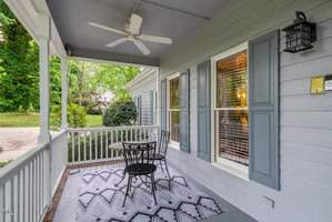 A cozy front porch with a black metal table and two chairs on a patterned rug. The light blue siding of the house features a window with white blinds. A ceiling fan hangs above, and trees are visible in the background.