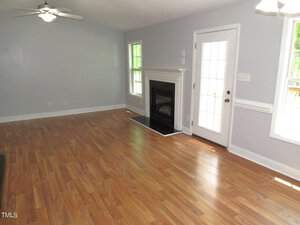 A bright living room with light gray walls, wooden laminate flooring, and a white ceiling fan. A fireplace with a white mantle is set between two tall windows. Near the back, a glass door leads outside, allowing natural light to enter the room.