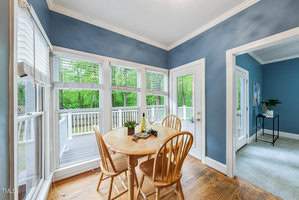 Bright dining area with wooden table and chairs set by large windows. Glass doors lead to a spacious deck. Room features blue walls and hardwood flooring. Outdoor view shows lush greenery.