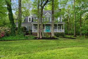 A charming two-story house with a light gray exterior and teal front doors is surrounded by lush green trees and well-maintained grass. The house features a wraparound porch and several large windows.