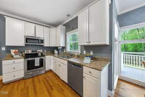 A modern kitchen with white cabinets, stainless steel appliances, and granite countertops. Wooden floors complement the space. Sunlight streams through a window and a glass door leading to a deck with a view of greenery outside.