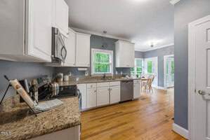A spacious kitchen with white cabinets, granite countertops, and stainless steel appliances. A window offers a view of greenery outside. A dining area with a table and chairs is visible in the background, near glass doors leading to a deck.