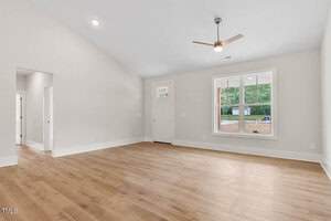 A spacious, empty room with light wood flooring and plain white walls. A large window lets in natural light, and a ceiling fan is centered on the high ceiling. A doorway leads to another room, and the front door is visible on the left.
