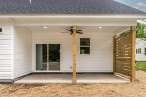 A covered white patio with a wooden privacy panel on the right and sliding glass doors leading inside. There's a ceiling fan and two recessed lights. The ground is dry with straw scattered around. Trees are visible in the distance.