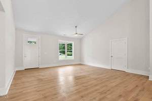 Spacious, empty living room with polished hardwood floor, beige walls, and high ceiling. Features a large window, ceiling fan, and a white front door. Natural light pours in from the window, highlighting the clean, minimalist design.