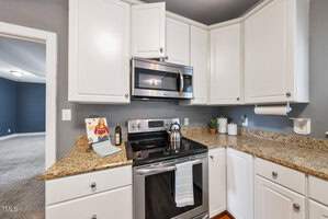 A modern kitchen with white cabinets, granite countertops, and stainless steel appliances, including a stove and microwave. A colorful cookbook, kettle, and small potted plant are on the counter. A doorway leads to a carpeted room.