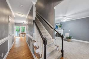 A modern home interior with a carpeted staircase leading to an upper floor. The living room features gray walls, a ceiling fan, and a fireplace. The hallway has wooden floors and leads to a dining area with large windows and a view of the outdoors.