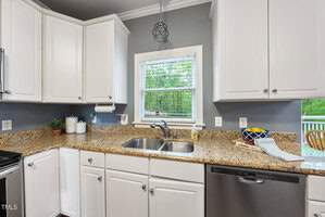 A modern kitchen with white cabinets, granite countertops, and a stainless steel sink. There is a window above the sink, a dishwasher beneath the counter, and a plant on the countertop. A geometric pendant light hangs from the ceiling.