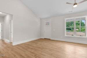 Spacious empty room with light wooden floors and white walls. A ceiling fan is mounted above, and sunlight streams through a large window to the right. A white door is near the window. The room has a high ceiling.