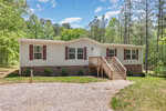 Located at 3206 Granville Ridge Road, this beige single-story house in Wake Forest features a brick foundation and brown shutters, nestled among trees. A wooden staircase leads to the front door, while a gravel driveway greets you in the foreground beneath a sky dotted with scattered clouds.