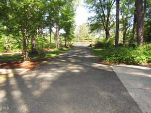 A gravel pathway leads through a lush, wooded area with tall trees on either side. Sunlight filters through the leaves, casting dappled shadows on the path. In the distance, a wooden fence is visible.