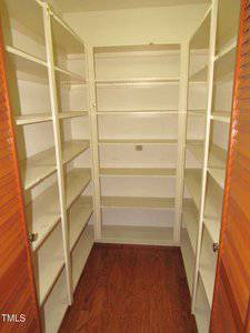 A small, empty pantry with wooden floors and white built-in shelves on three walls. The pantry is enclosed by orange louvered doors.