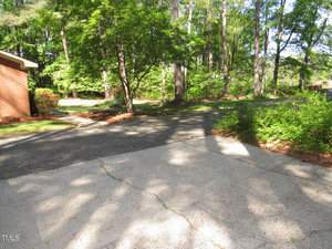 A sunlit driveway surrounded by lush green trees and bushes. The paved path curves past a red-brick building on the left, leading into a wooded area with tall trees in the background. Shadows of trees are cast across the concrete surface.