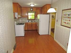 A small kitchen with wooden cabinets, a white gas stove, and a refrigerator. The room features a hardwood floor and floral wallpaper. A window above the sink provides natural light, and a framed vegetable alphabet poster hangs on the wall.